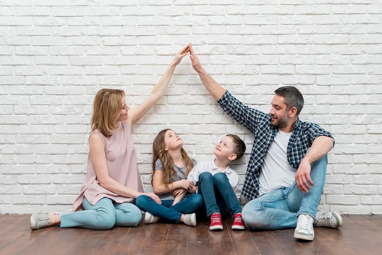 Beyaz tuğla duvar önünde yerde oturan anne, baba ve iki çocuk; anne ile baba ellerini birleştirerek çocukların üzerinde çatı şekli oluşturuyor.